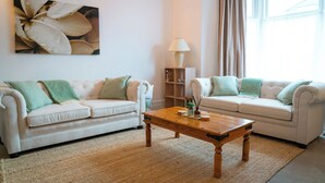 Un salon avec deux canapés blancs, une table basse en bois, une lampe et un grand tableau floral accroché au mur.