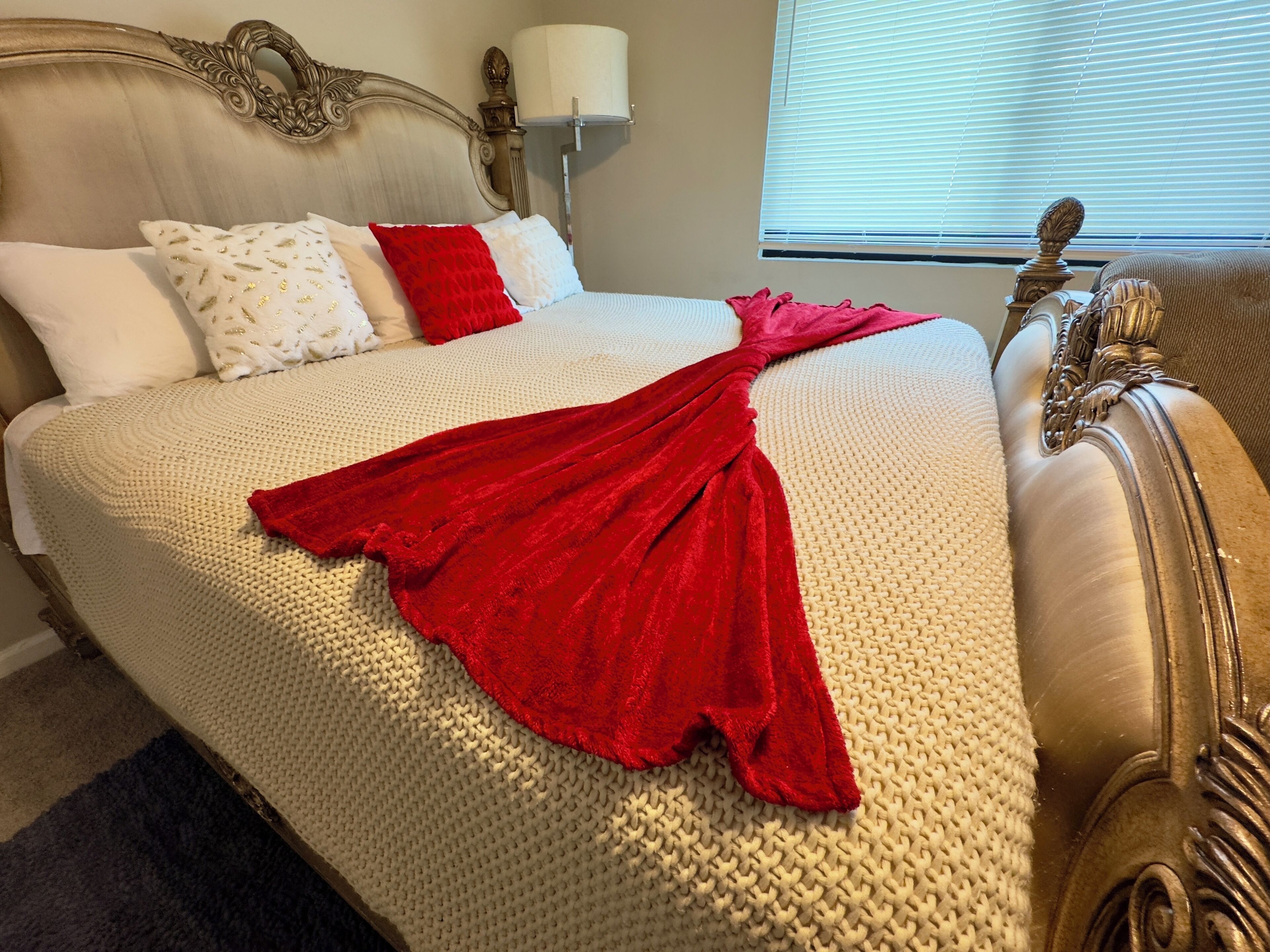 Una cama con cubierta de punto color beige, una manta roja y cojines decorativos.