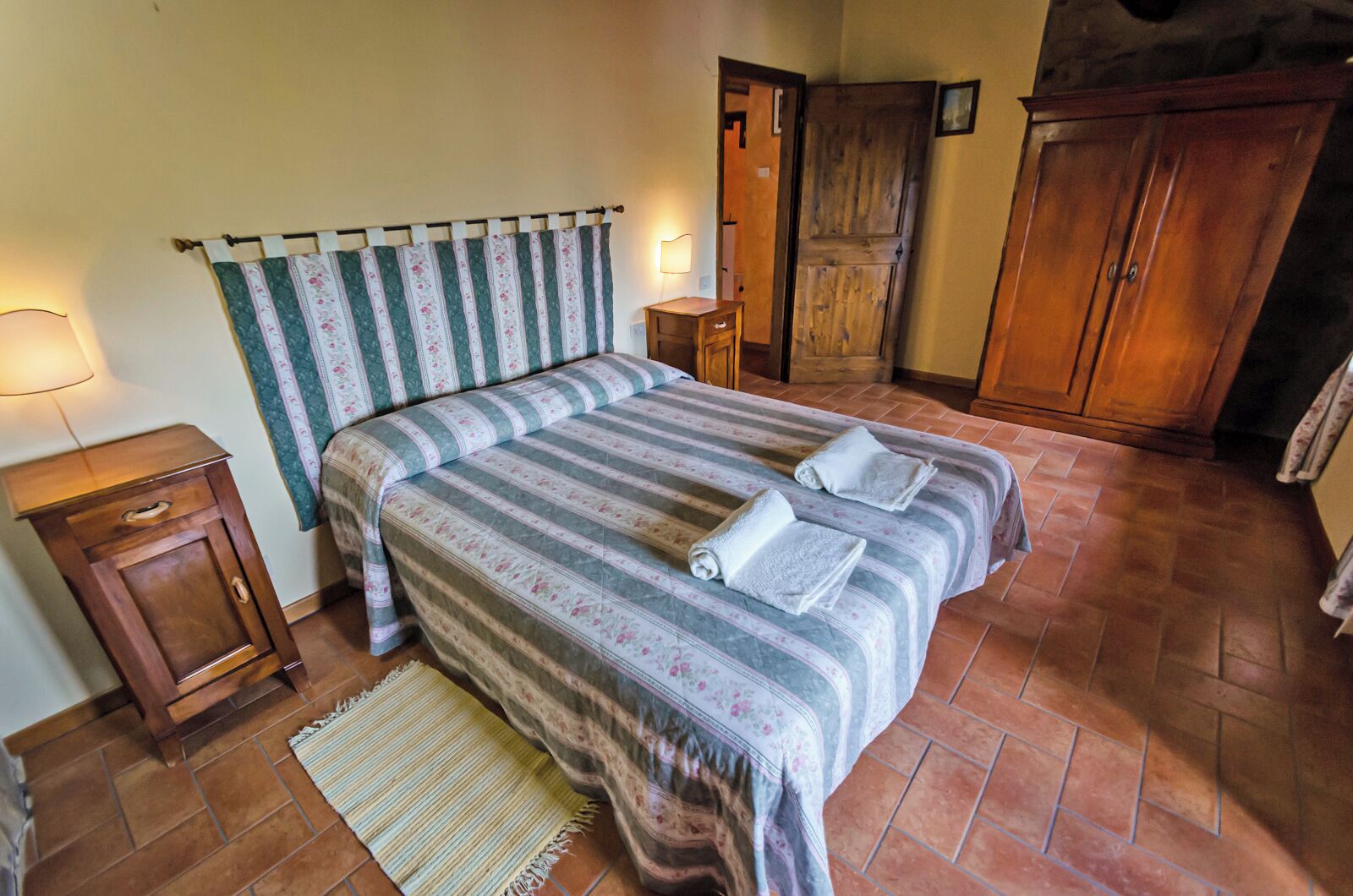 A bedroom with a bed, two nightstands, a lamp, a wooden wardrobe, and a door.