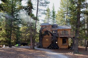 Una cabaña tipo casa del árbol con fachada de madera, construida en un árbol grande, que cuenta con un balcón y una ventana pequeña.