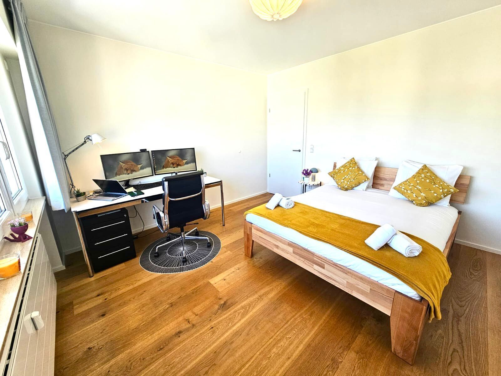A bedroom with a bed, a desk with two monitors, a chair, and a small rug.