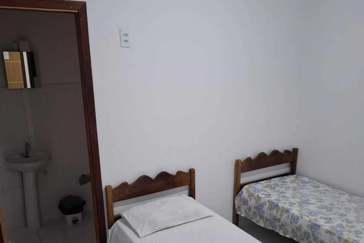Quarto pequeno e limpo, com duas camas de solteiro, cabeceira de madeira, parede branca e banheiro pequeno com pia e vaso sanitário.