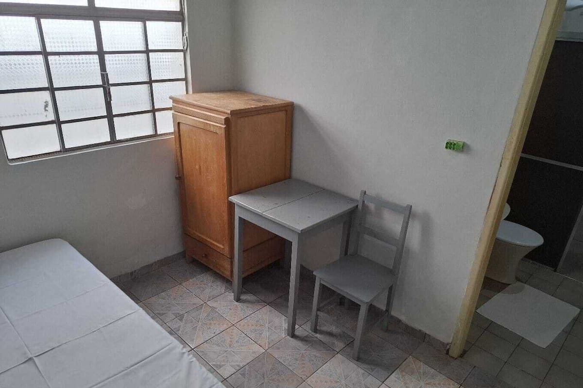 Um quarto pequeno e pouco mobiliado, com uma cama, um armário de madeira, uma mesinha e uma cadeira.