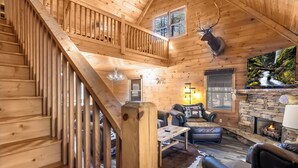 L'intérieur d'un chalet en bois, avec une cheminée en pierre, des meubles en cuir, une tête de cerf fixée au mur et un téléviseur diffusant une scène de cascade.