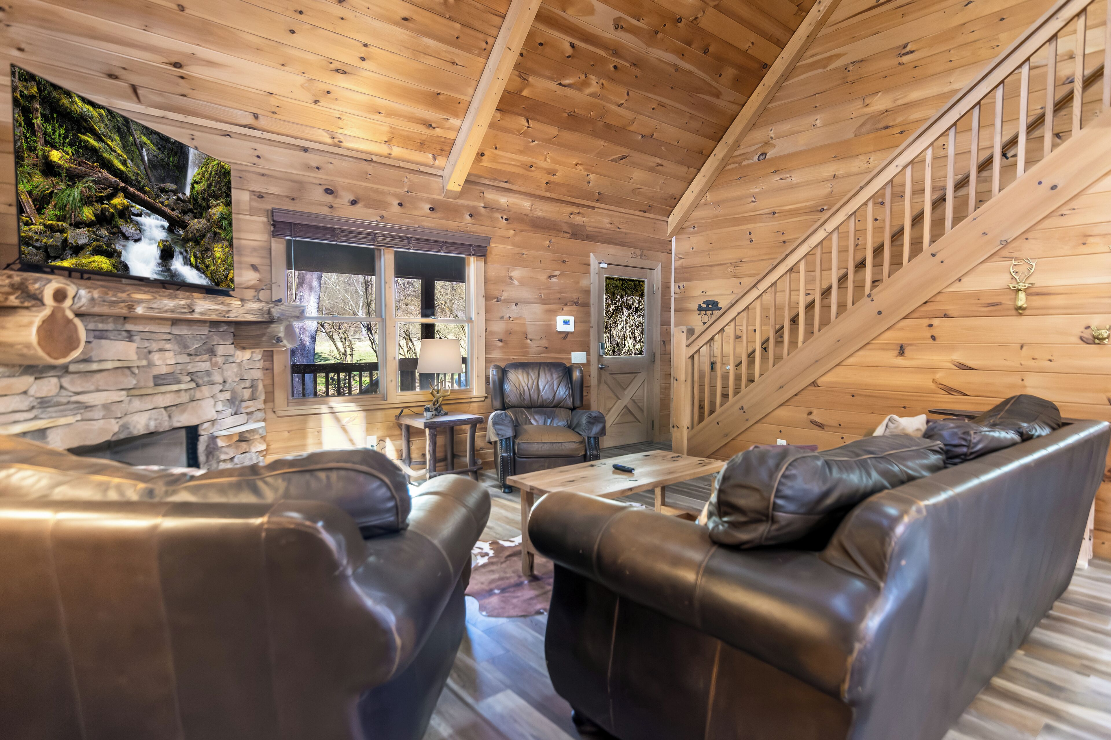 L’intérieur chaleureux d’un chalet avec une cheminée en pierre, des canapés en cuir, un escalier en bois et un grand téléviseur diffusant des images de la nature.