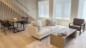 A modern living room with a sofa, coffee table, dining table, and chairs. There is a staircase with a wooden railing and a window with blinds. - 2BR 2.5 Downtown Oasis Full Kitchen, Pet Friendly, Close to Silos Food Hall (Easley)
