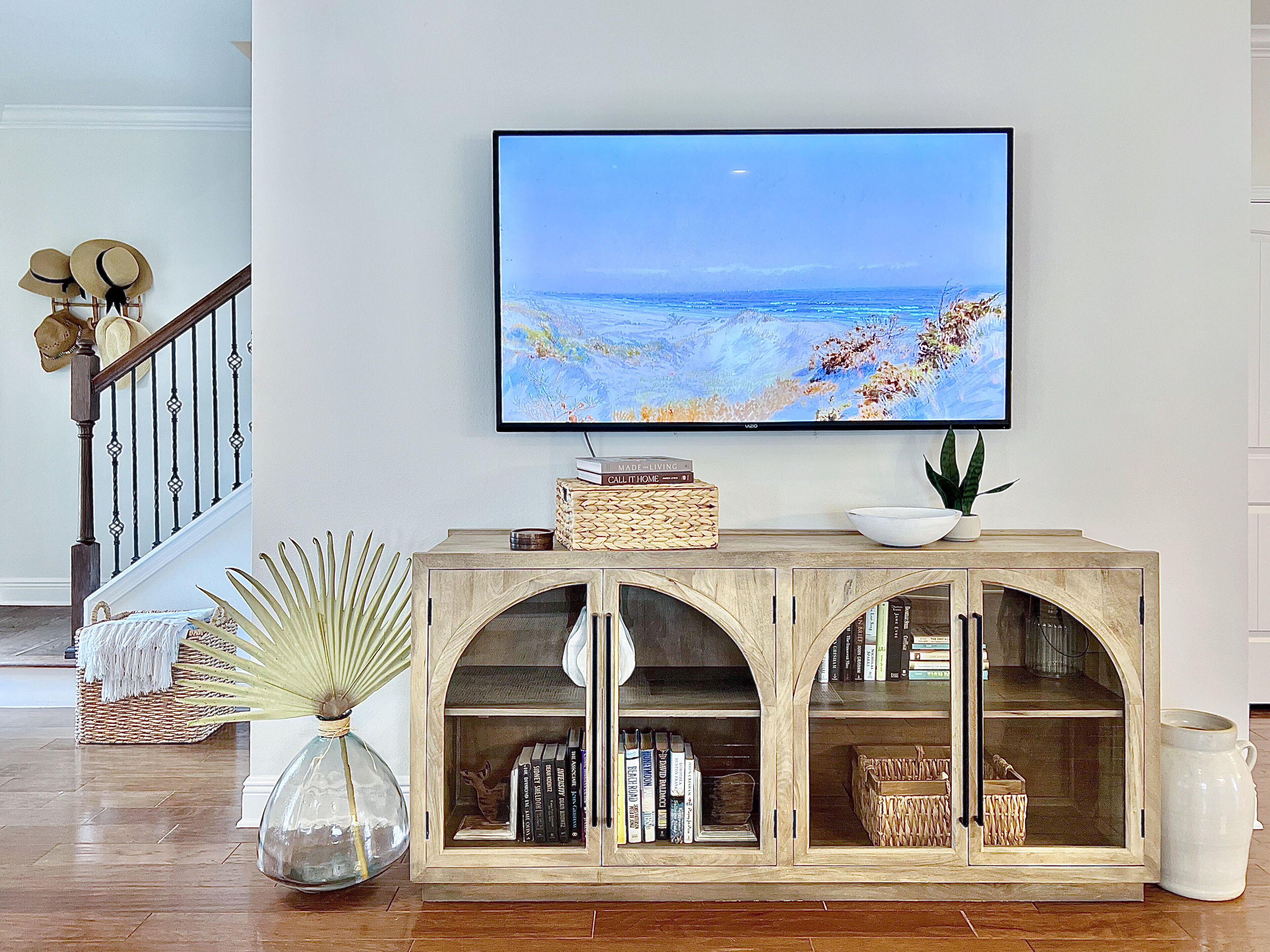 Uma sala de estar com televisão na parede, um console de madeira com livros e objetos decorativos, e uma escada com corrimão.