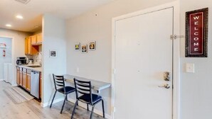Une cuisine compacte avec des meubles en bois, un four encastré et un coin repas avec deux chaises. Un panneau indiquant « BIENVENUE » est accroché au mur.
