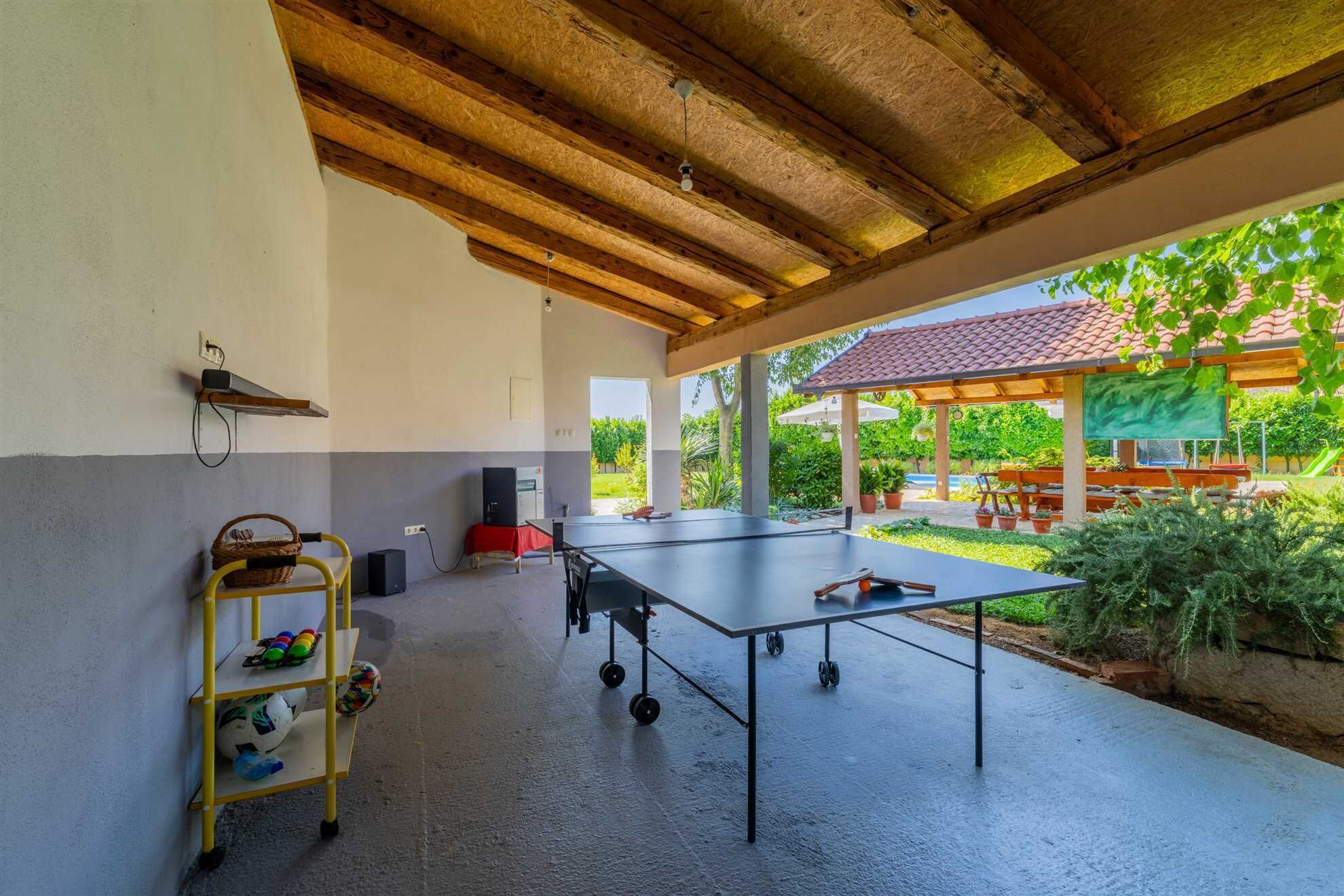 A covered outdoor area with a ping pong table, a yellow shelf with sports equipment, and a view of a garden and outdoor seating area.