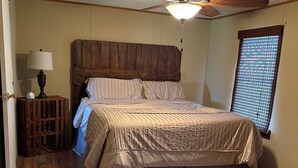 A bedroom with a wooden headboard, a bed with striped bedding, a floor lamp, a window with blinds, and a ceiling fan.