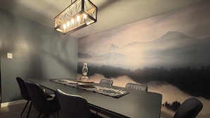 A dining room with a long table, six chairs, a chandelier, and a wall mural of a mountain landscape.