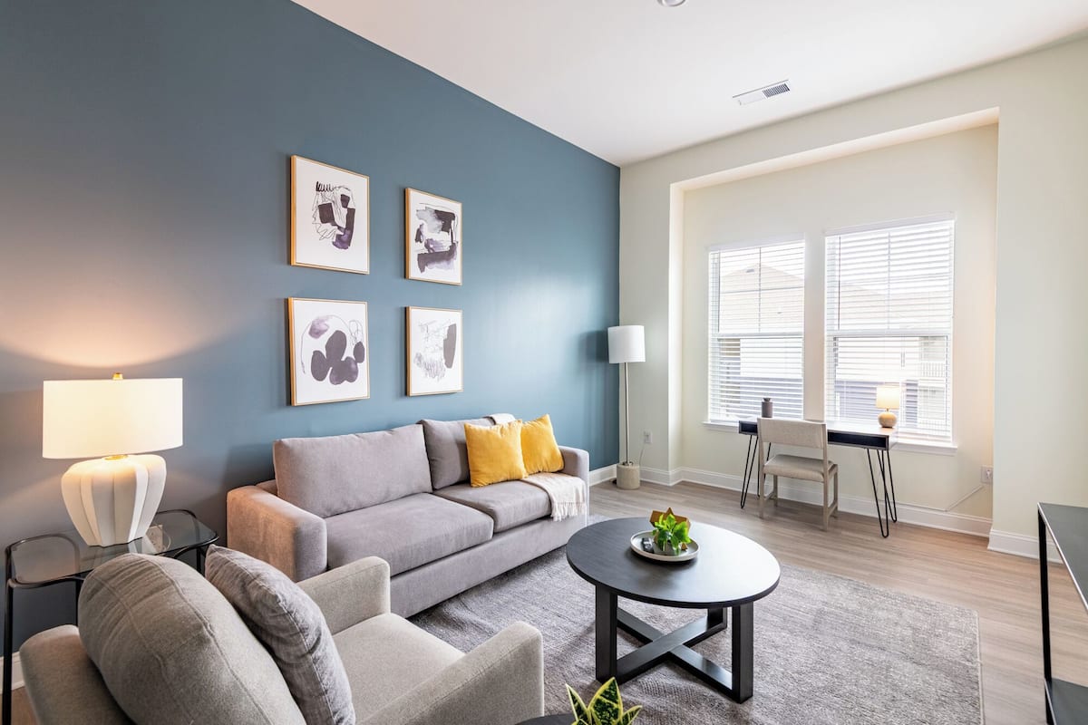 Ein modernes Wohnzimmer mit grauem Sofa, rundem Couchtisch und einer blauen Akzentwand. Vier gerahmte Kunstwerke sind an der Wand angebracht, und auf einem Beistelltisch steht eine Lampe.