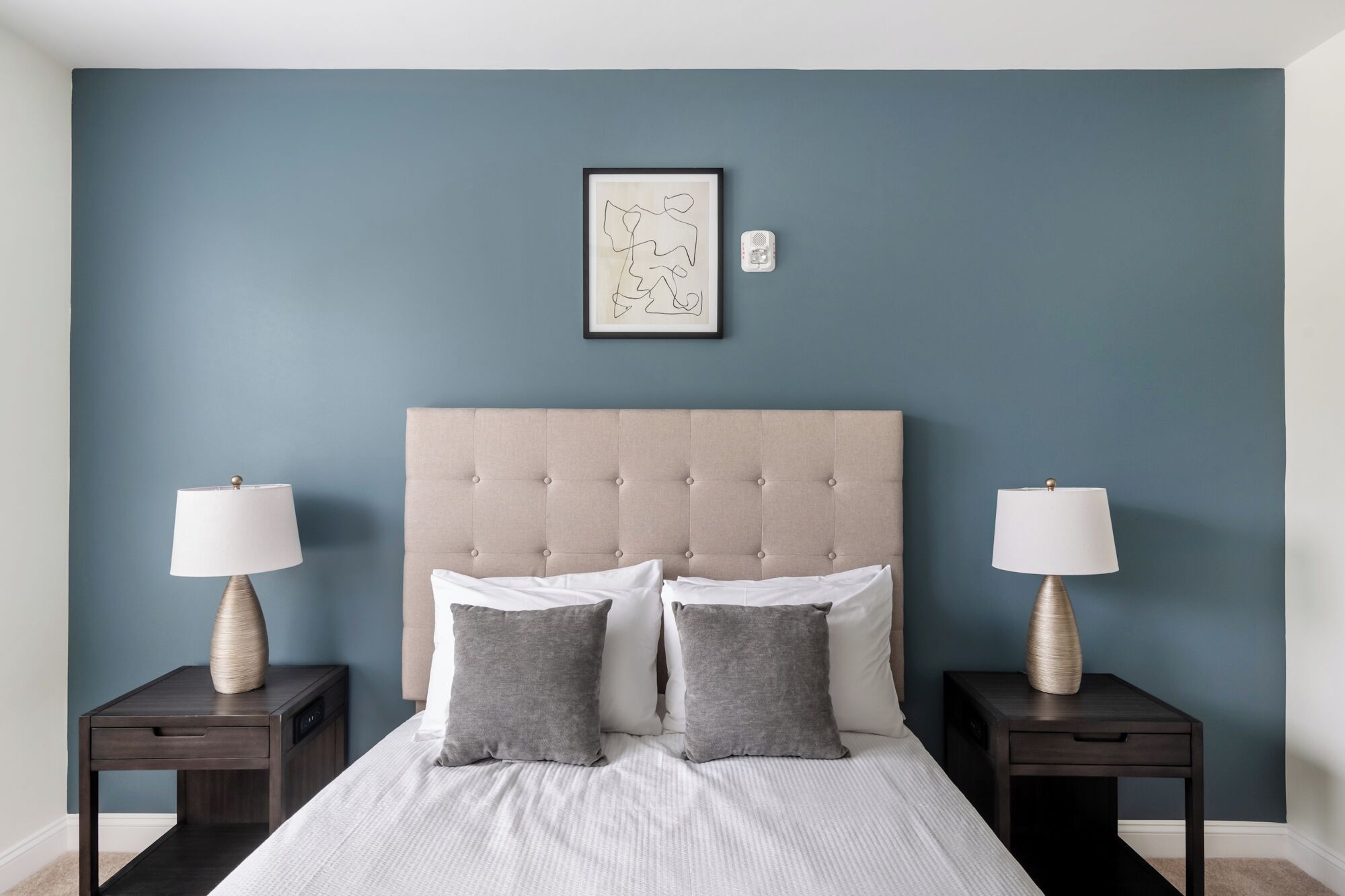 Une chambre à coucher avec une tête de lit capitonnée, un lit recouvert de draps blancs et agrémenté de deux oreillers gris, deux tables de chevet avec des lampes, et une œuvre d’art abstraque encadrée accrochée au mur.