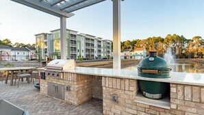 An outdoor kitchen area with a grill, a large green smoker, and a stone countertop. - Landing Apartments Santa Rosa Beach (Santa Rosa Beach)