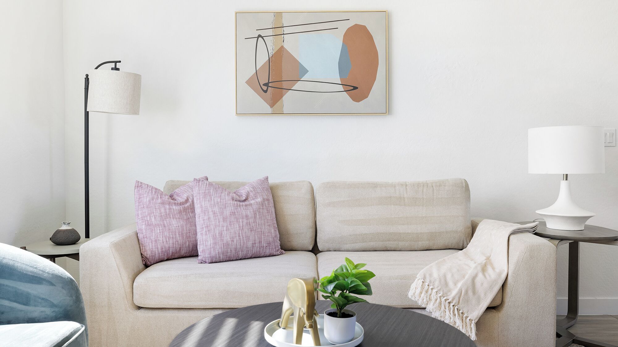 Un soggiorno con un divano beige, due cuscini, una piccola pianta in vaso e una lampada su un tavolino.