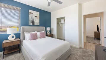 Un dormitorio con cama, mesitas de noche, ventilador de techo, ventana con persianas y un cuadro en la pared.