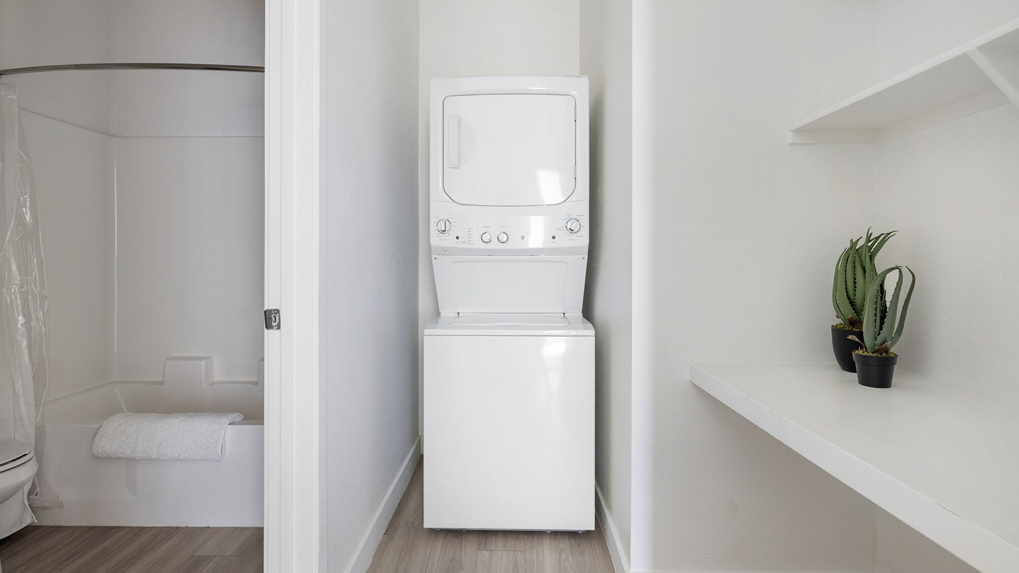 Une petite buanderie comprenant un lave-linge et un sèche-linge, un espace douche et une petite étagère avec des plantes en pot.