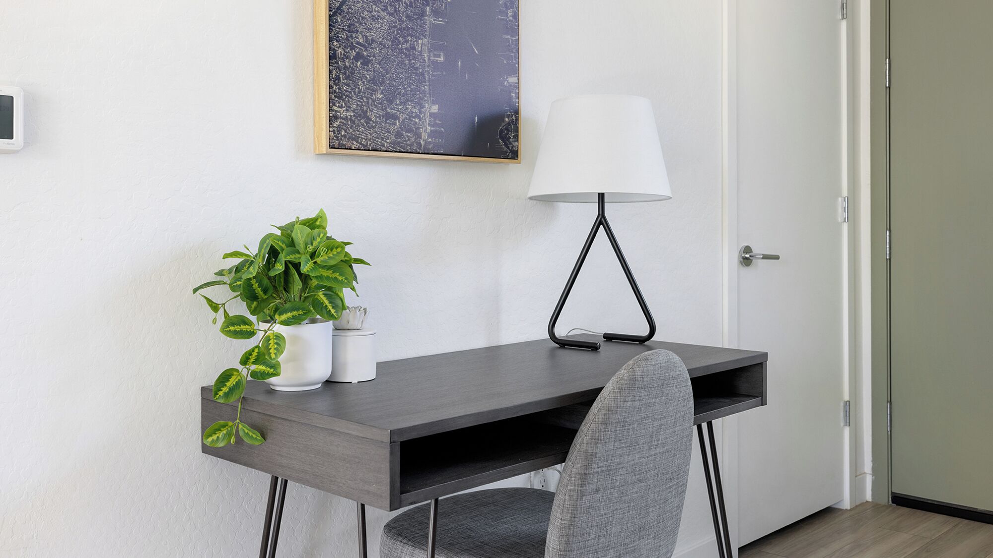 A modern desk with a plant, a lamp, and a cityscape painting on the wall.