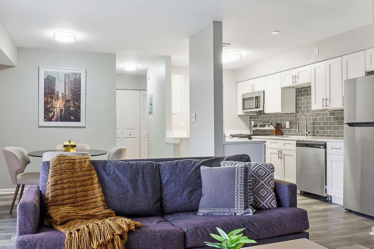Un appartement moderne doté d’un salon avec un canapé violet, d’un coin repas avec une petite table et d’une cuisine avec des meubles blancs et des appareils en acier inoxydable.