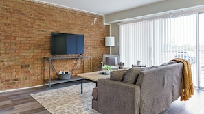 Ein Wohnzimmer mit Backsteinwand, einem Flachbildfernseher an einer Metallhalterung, einem grauen Sofa, einem kleinen Tisch mit Pflanze und einem großen Fenster mit Jalousien.