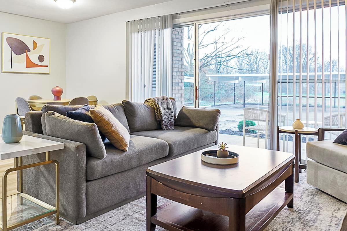 Un salon moderne avec un canapé gris, une table basse et une table d’appoint. Un tableau est accroché au mur et il y a une fenêtre avec des stores.