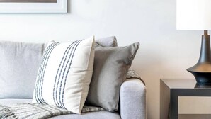 Ein graues Sofa mit zwei Kissen, eines gestreift, eines schlicht, und eine schwarze Lampe auf einem kleinen Tisch daneben.