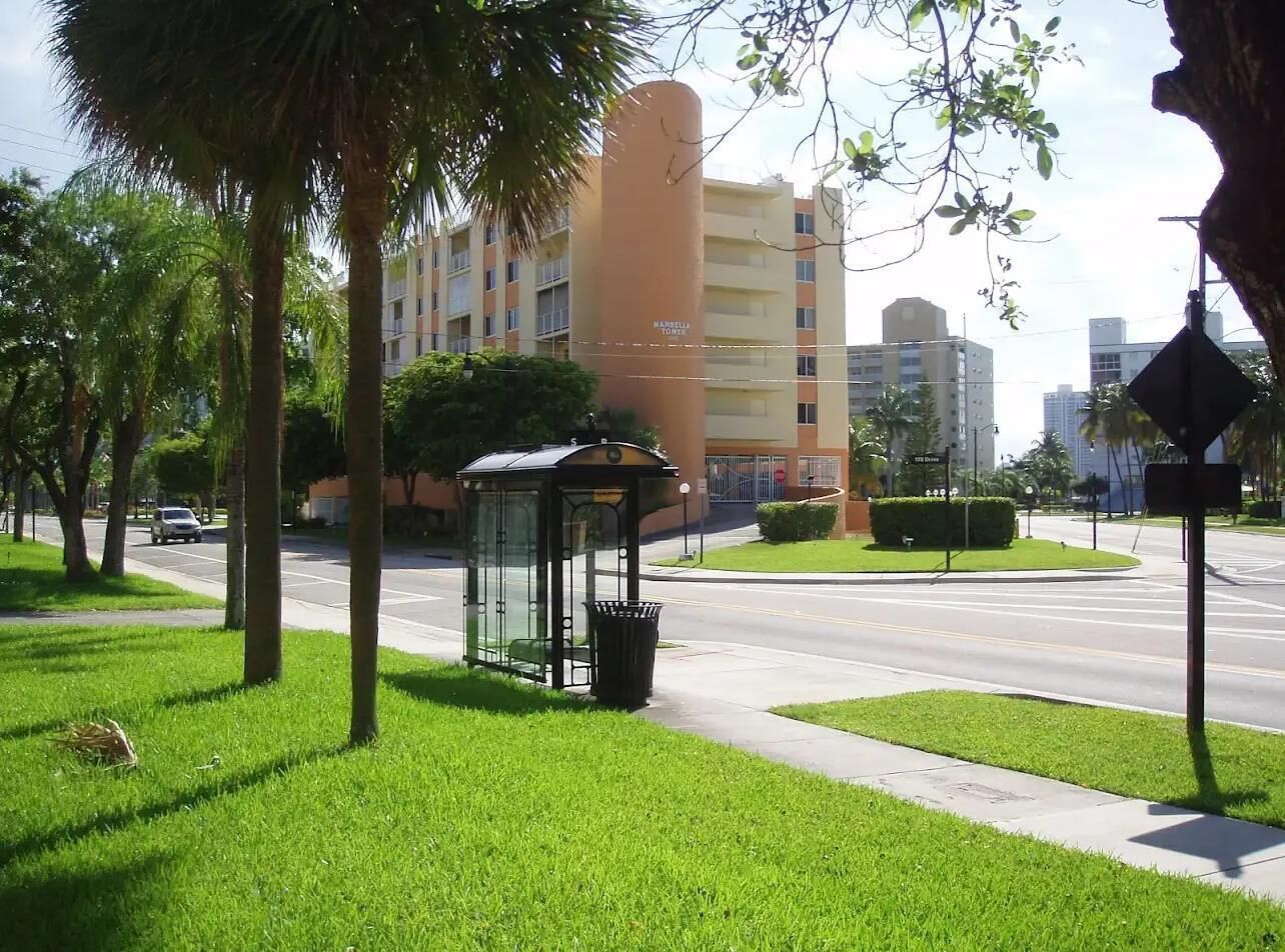 Una calle urbana verde con una parada de autobús, un cubo de basura y un edificio al fondo.