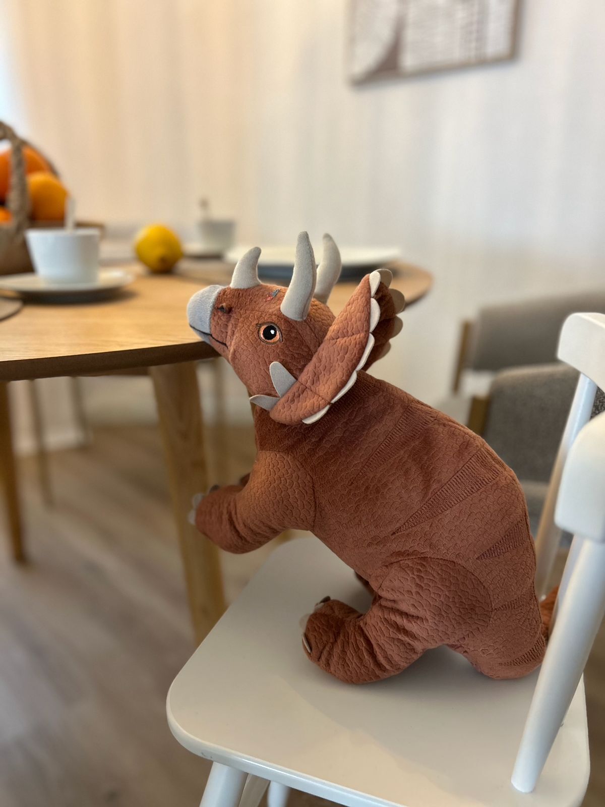 Ein weiches Plüschdinosaurier sitzt auf einem weißen Stuhl in einem Raum mit einem Holztisch und einer weißen Wand.