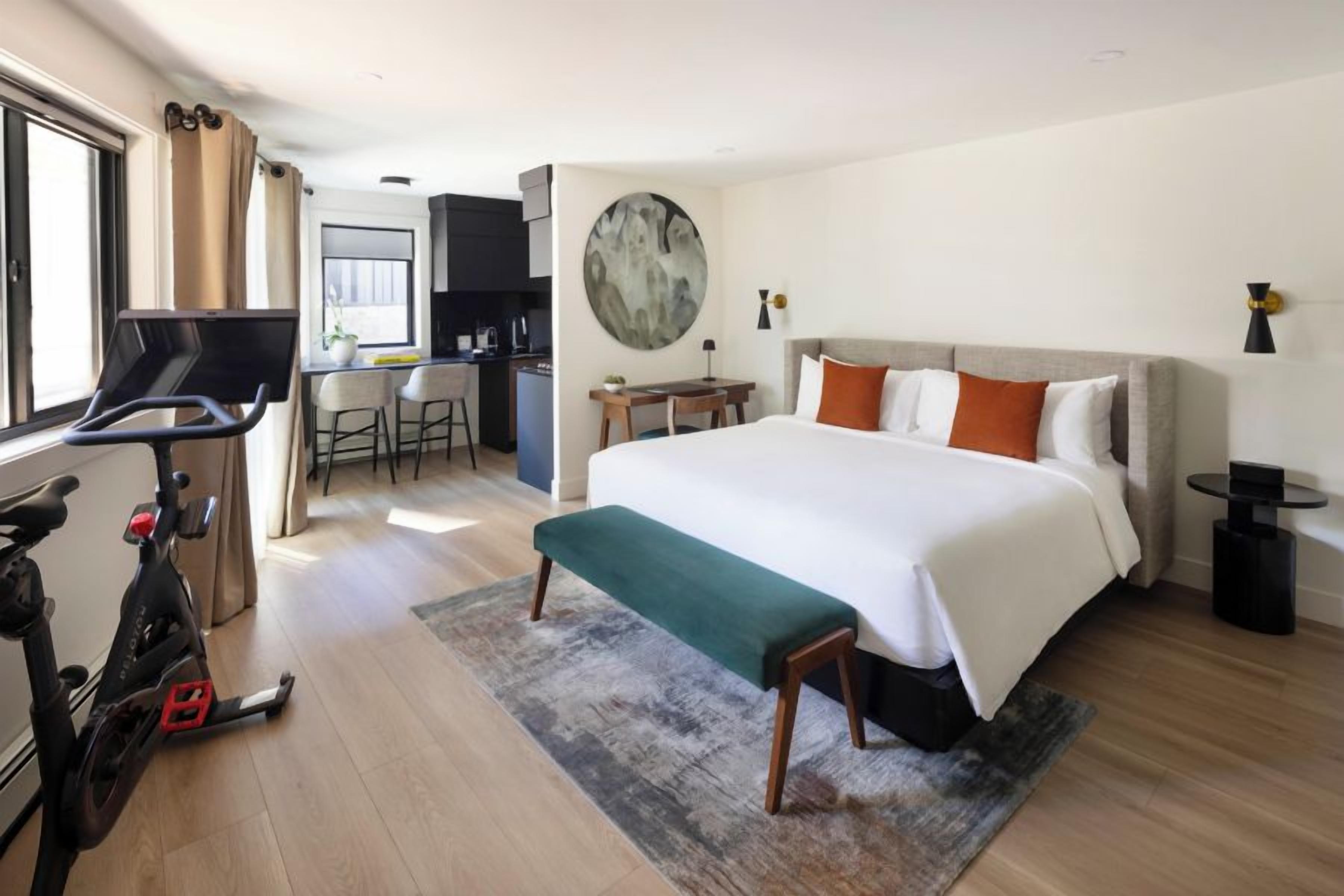 A hotel room with a bed, a desk, a chair, a treadmill, and a large window.
