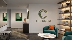 Um interior moderno com uma mesa de recepção, uma área de estar com uma poltrona e apoio para os pés verde-água, uma mesa de centro de mármore e uma estante com objetos decorativos.