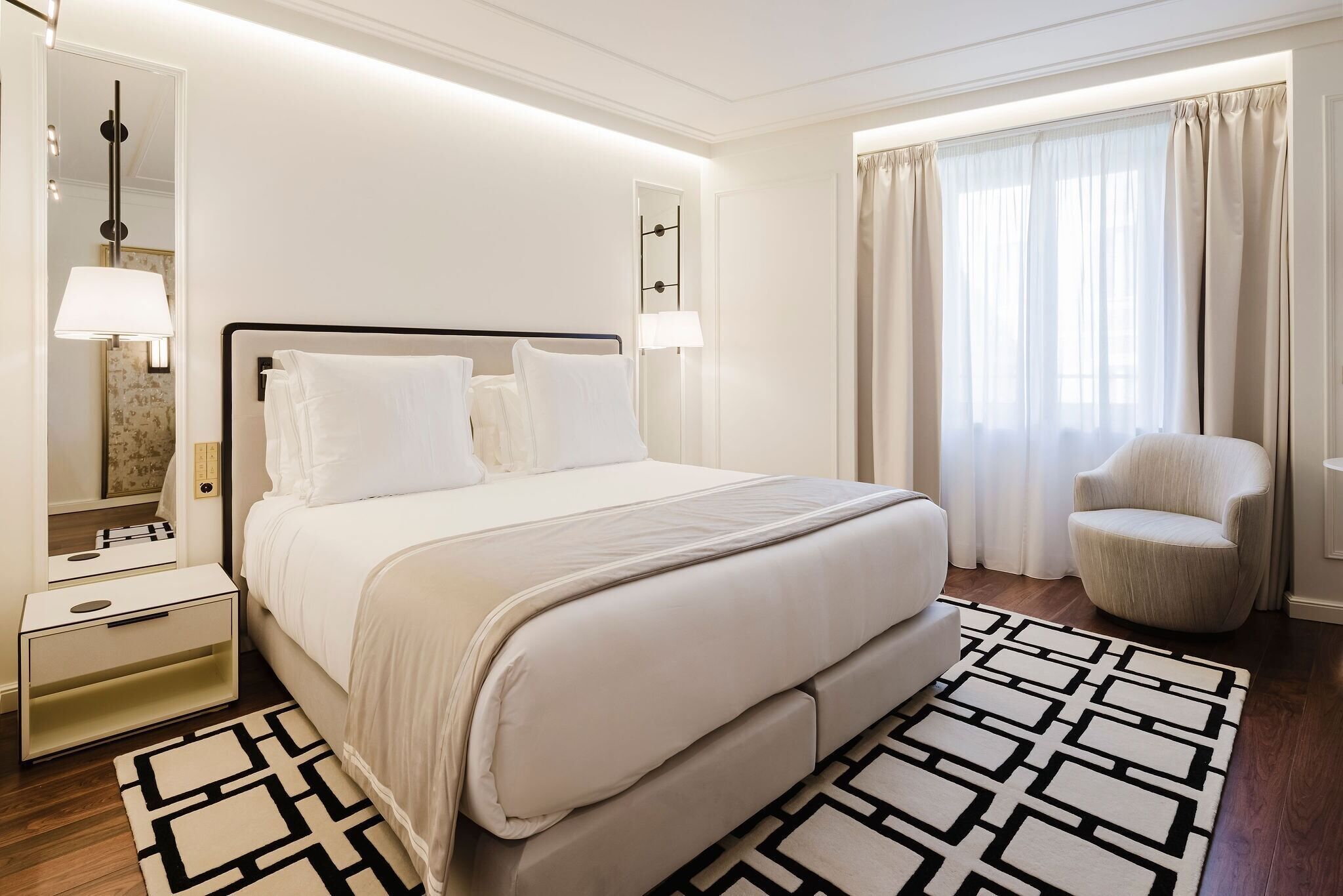Una habitación de hotel con cama, mesitas de noche, una silla, un espejo y una ventana con cortinas.