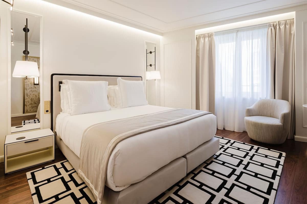 Una camera d'albergo con un letto, comodini, una sedia, uno specchio e una finestra con tende.