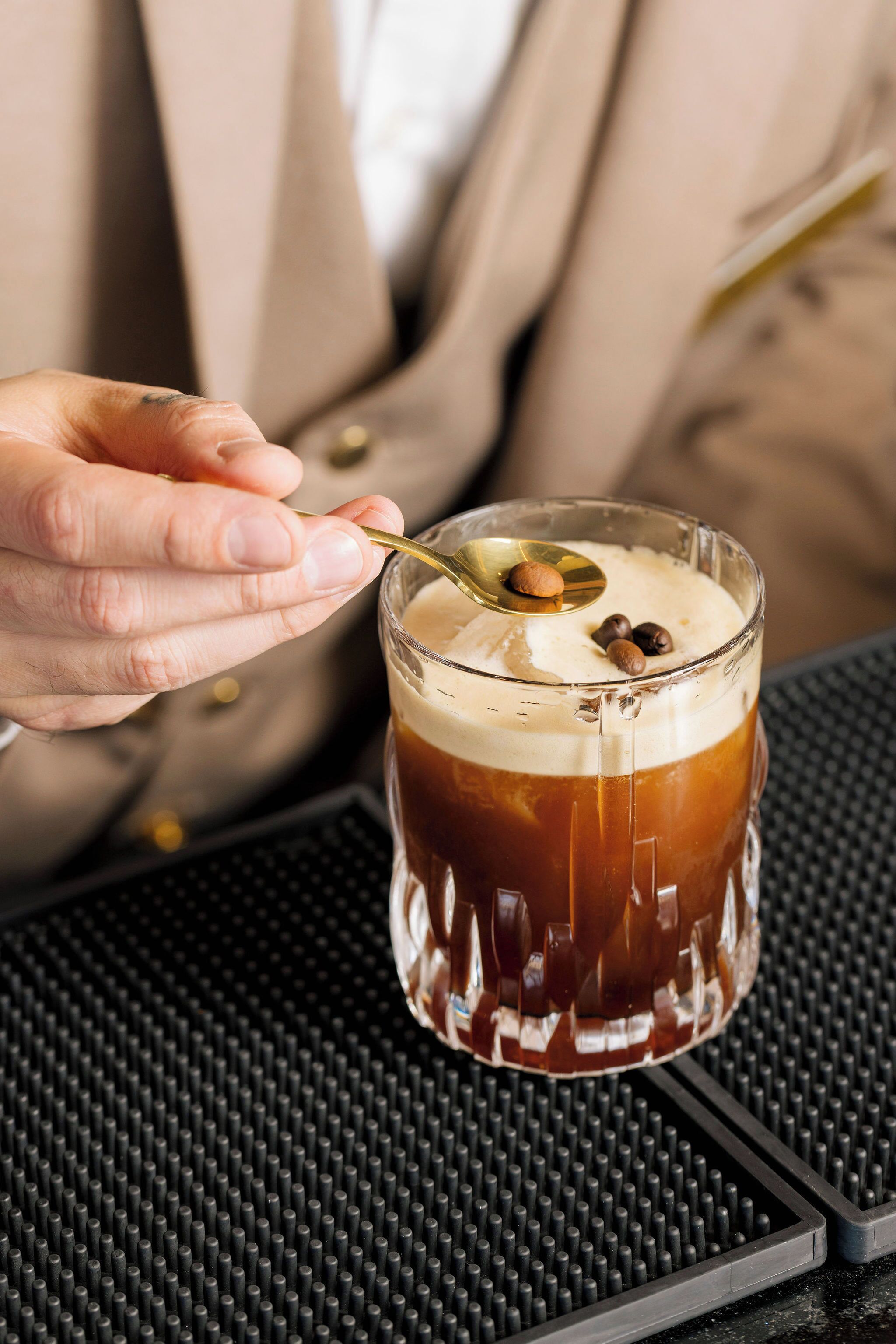 A hand holding a glass with a spoon, containing a beverage with coffee and chocolate pieces.