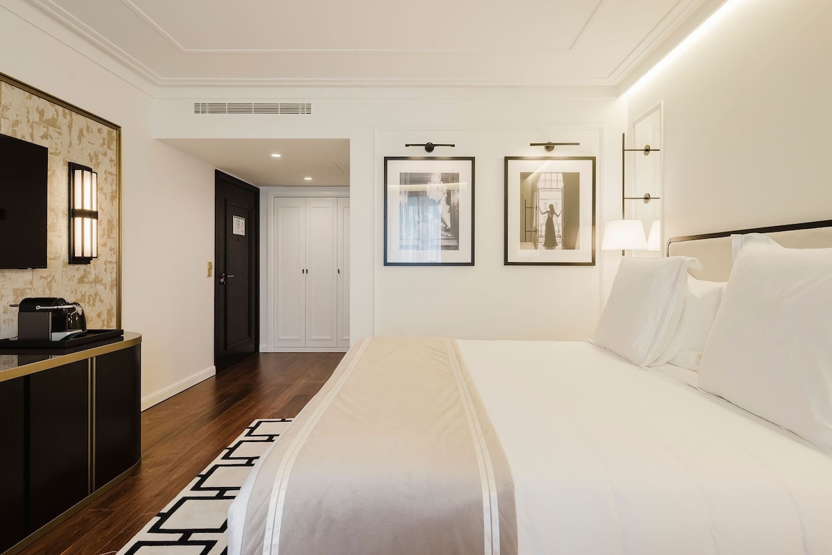 Hotelkamer met een bed, bureau, televisie, ingelijste foto’s aan de muur en een deur naar een andere kamer.