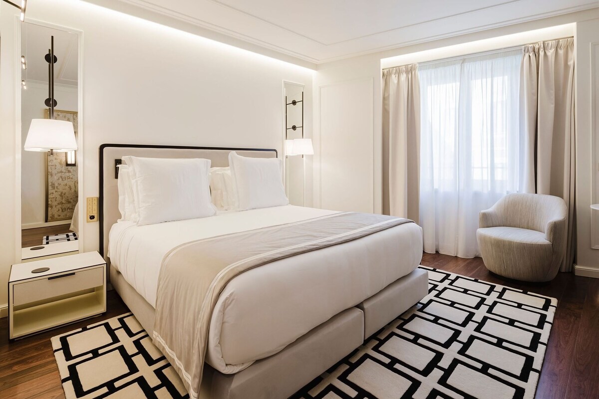 Une chambre d’hôtel avec un lit, des tables de chevet, une chaise, un miroir et une fenêtre avec des rideaux.
