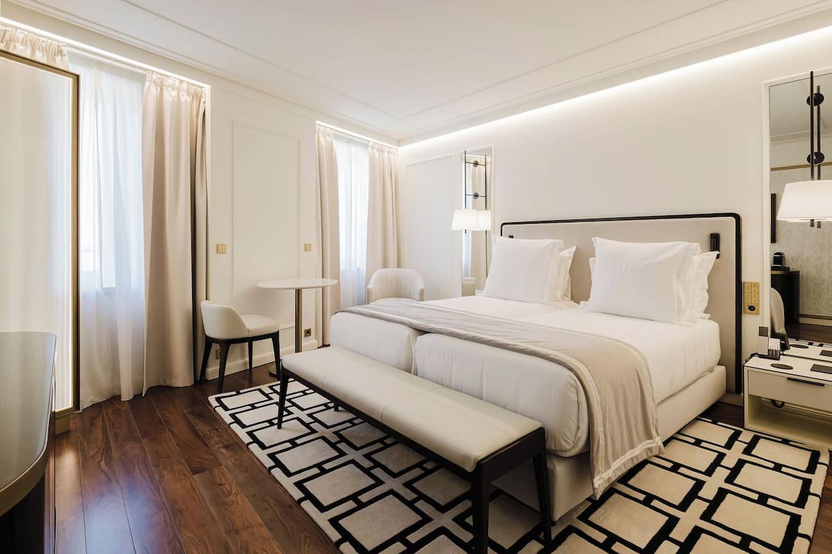 A hotel room with a large bed, a bedside table, a chair, a bench, and a patterned rug.