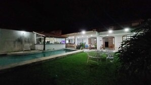 Vista nocturna de una propiedad residencial con piscina, césped y zona de comedor con mobiliario exterior.