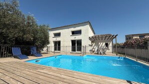 Ein Schwimmbecken mit Holzterrasse, ein Gebäude mit einer Pergola und ein Zaun.