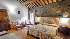 Un dormitorio con pared de piedra, vigas de madera, cama con ropa de cama floral, una silla, una mesa y una pequeña estatua.