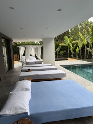 Un espace piscine extérieur moderne, doté de trois chaises longues et d’un lit à baldaquin, le tout entouré d’une végétation luxuriante.