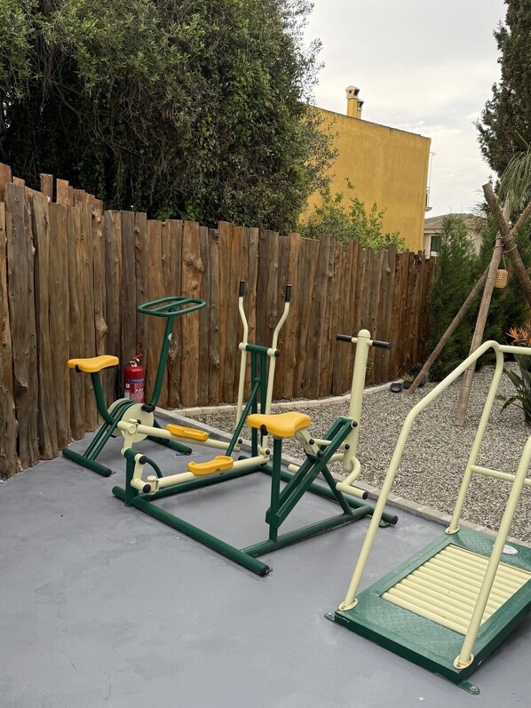 Outdoor exercise equipment, including a stepper and a bench, set against a wooden fence and a building in the background.