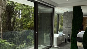 A modern bedroom with a large bed, a glass door leading to a balcony, and a view of a forest.
