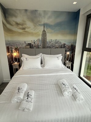 Une chambre d’hôtel avec un grand lit, une fresque représentant un paysage urbain sur le mur, et deux tables de chevet avec des lampes.