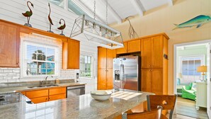 A kitchen with wooden cabinets, a granite countertop, a stainless steel refrigerator, and a dining area with chairs. - Cottage on the Canal with Tiki Bar – Relax & Unwind in Ocean Isle (Ocean Isle Beach)