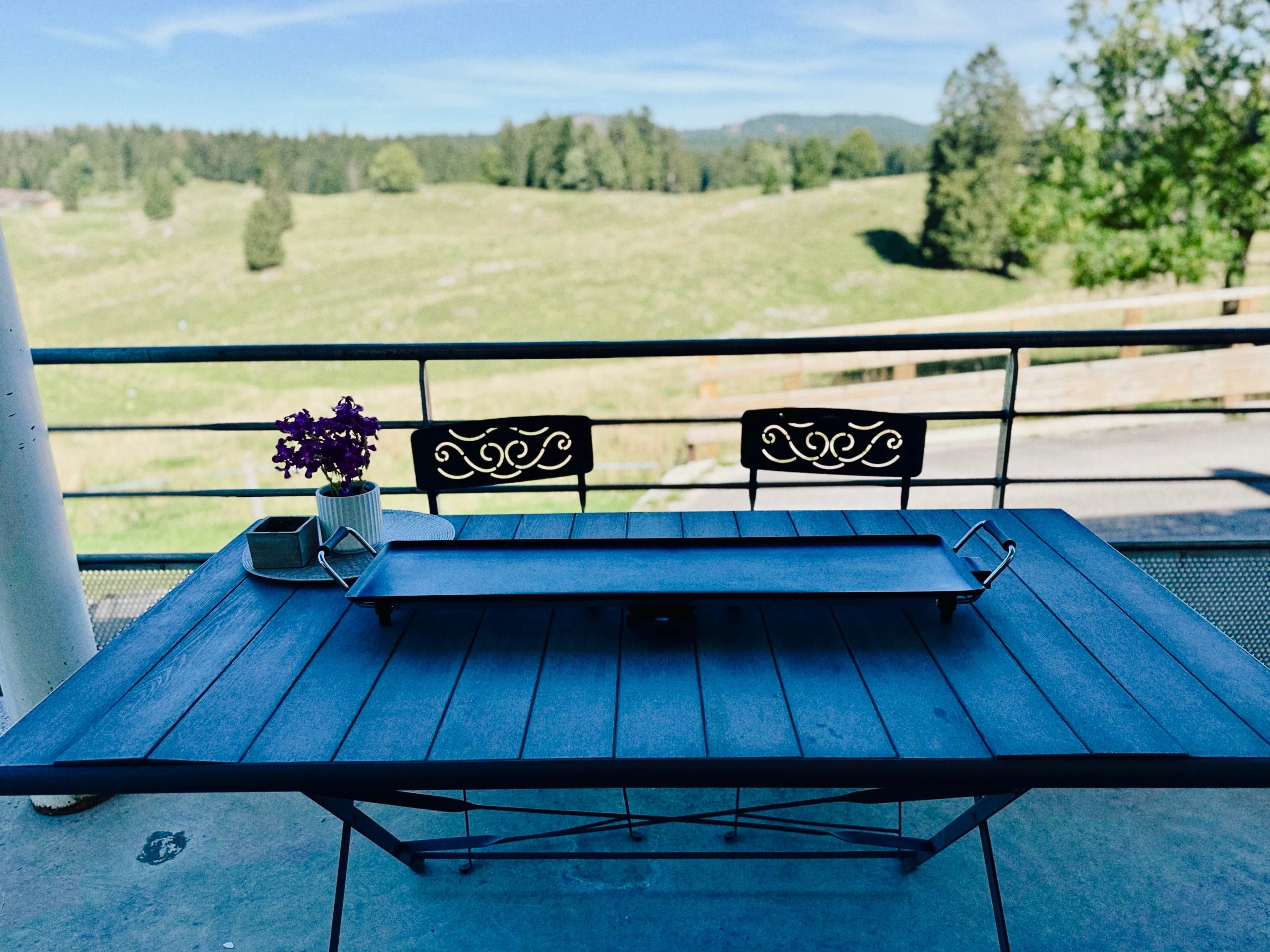 Un balcon avec une table bleue et deux chaises, un pot de fleurs et une vue sur un paysage verdoyant.