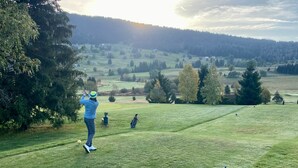 Un terrain de golf avec une personne qui s’apprête à frapper une balle, un seau bleu et une voiturette de golf.