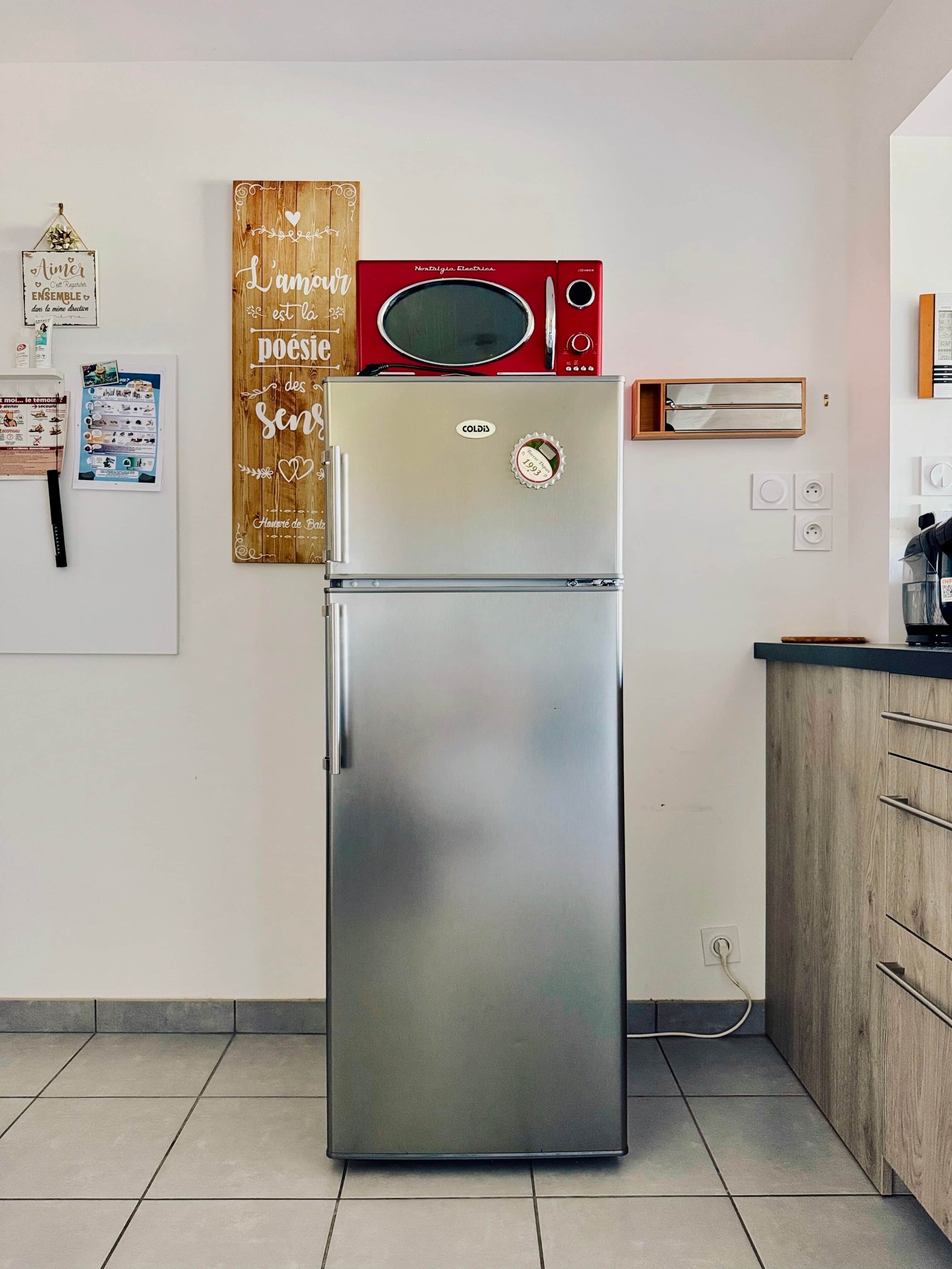 Ein Edelstahl-Kühlschrank mit Gefrierfach, darüber eine Mikrowelle und ein hölzernes Schild mit Schriftzug an der Wand.