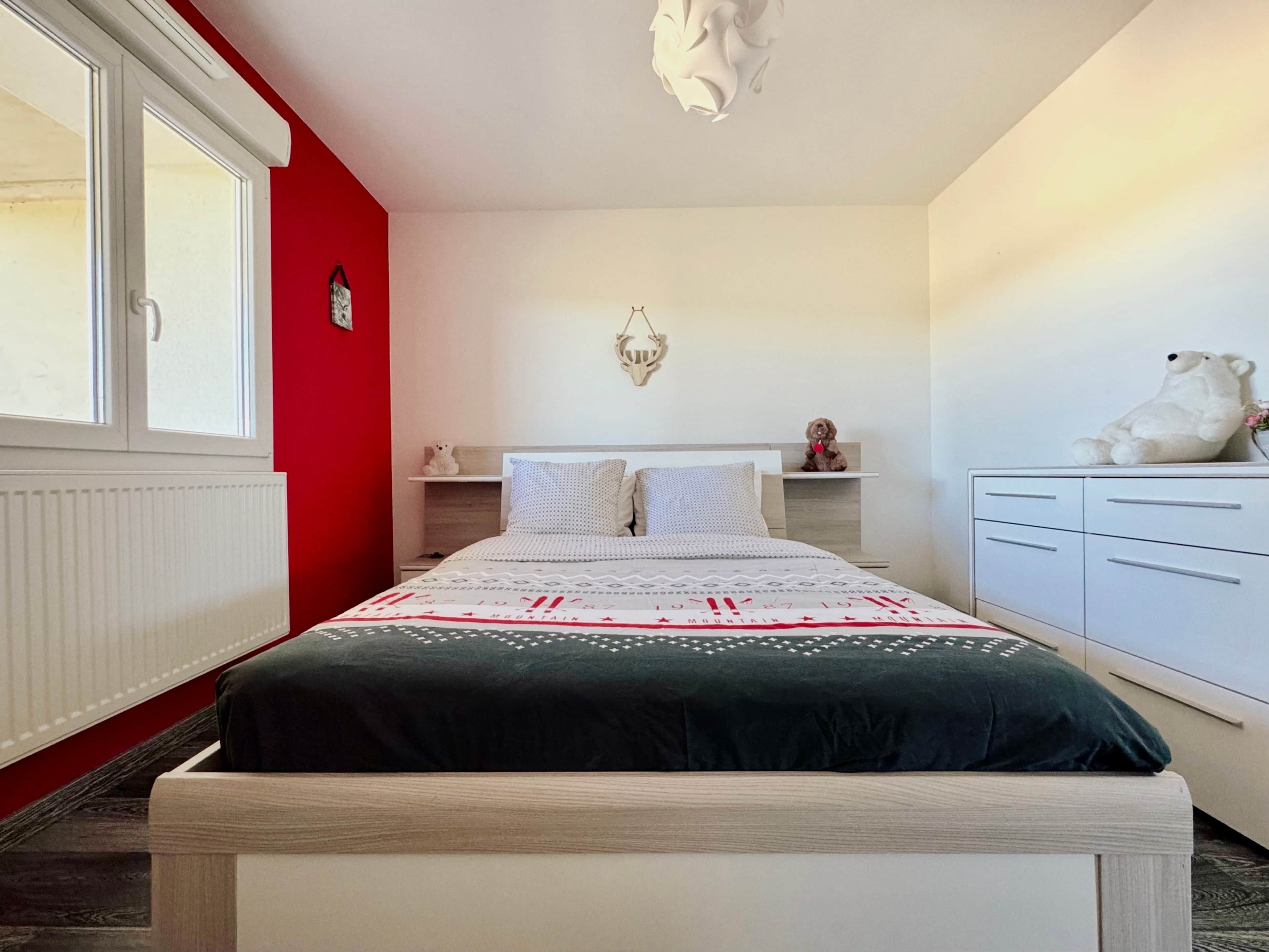 A bedroom with a bed, a dresser, a nightstand, a window, and a wall-mounted light fixture.
