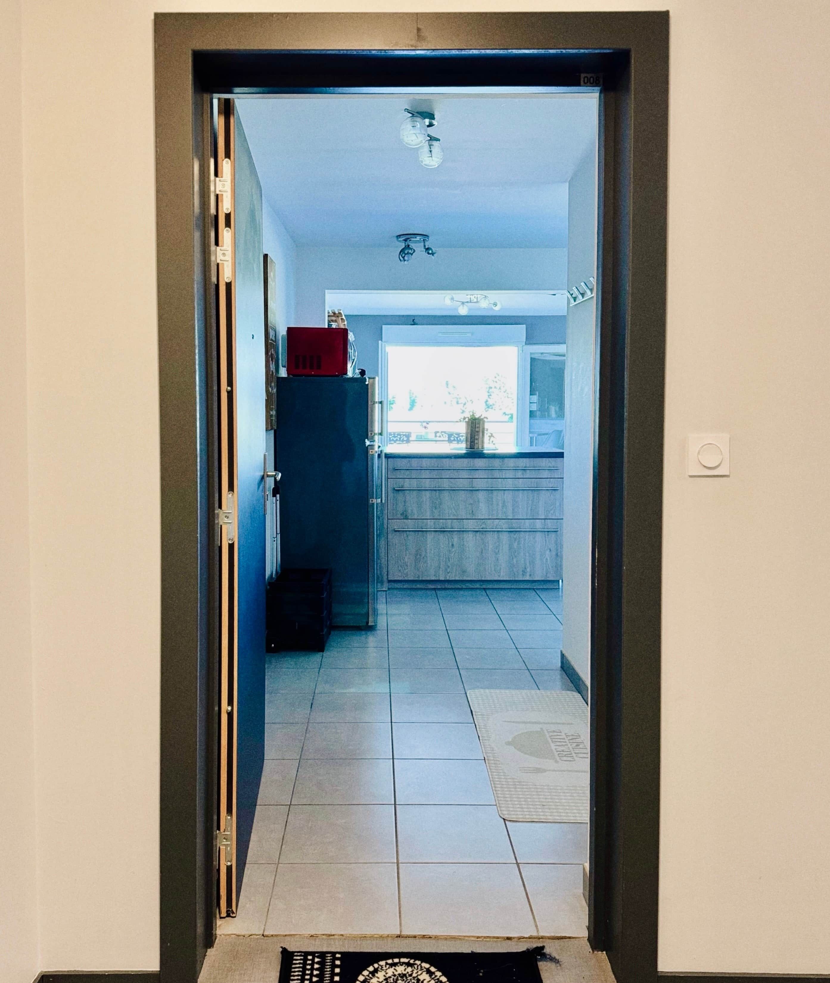 Un couloir mène à une cuisine dotée d’un sol carrelé, d’un réfrigérateur et d’une fenêtre offrant une vue agréable.
