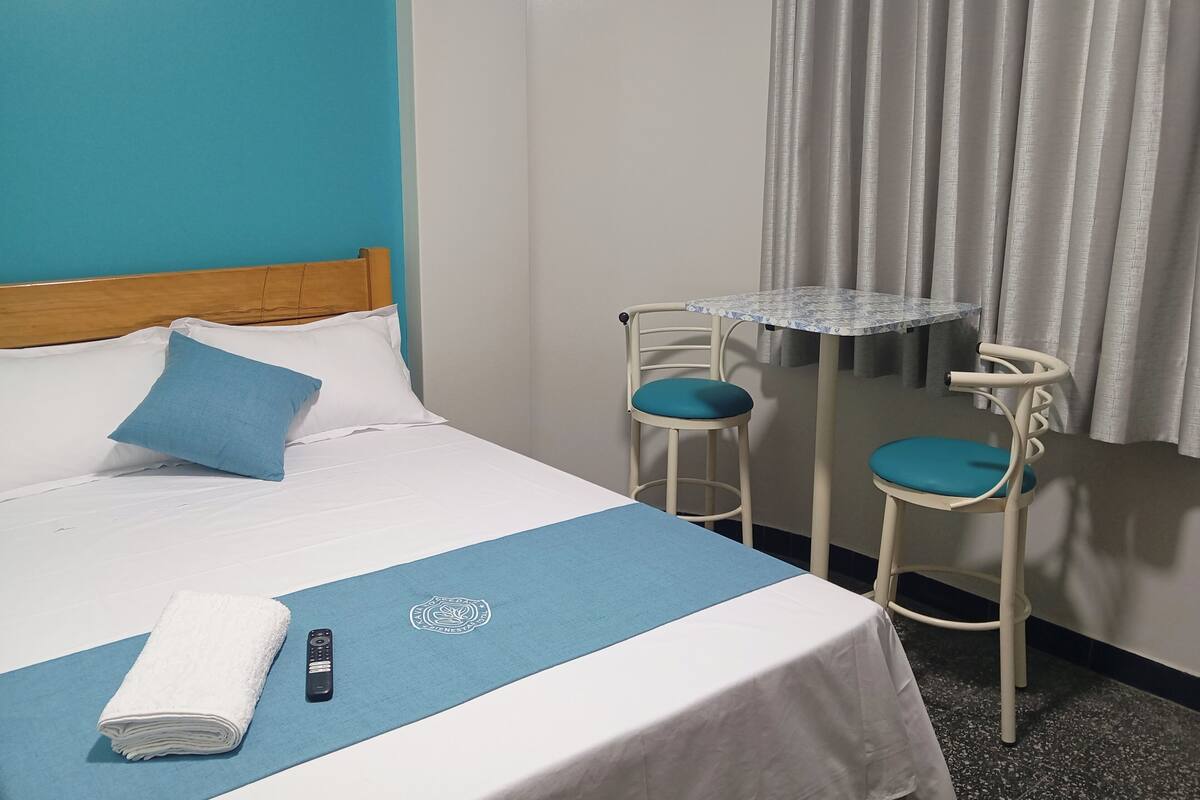 Una cama bien hecha con un almohadón azul y una manta blanca, una mesita con una silla azul, y un mando a distancia sobre la cama.